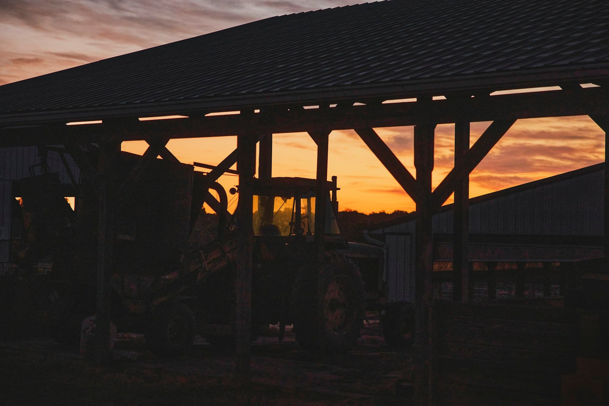 A tractor silhouetted under a barn roof against an orange sunset sky
