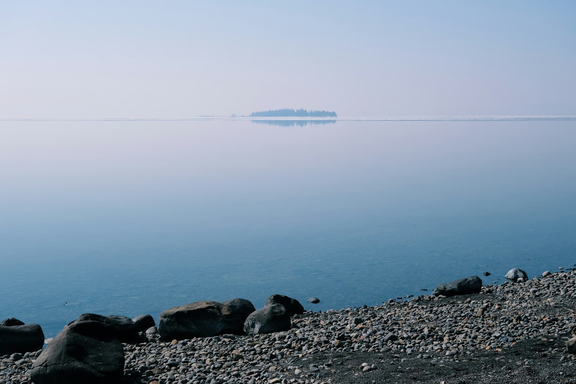 A still blue lake in soft haze with a distant island and a rocky pebble shoreline