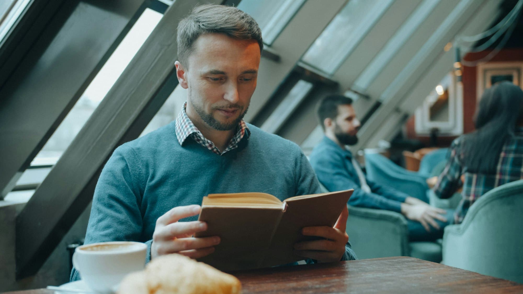 A man reads a plain-covered book at a cafe table beside a coffee and a croissant