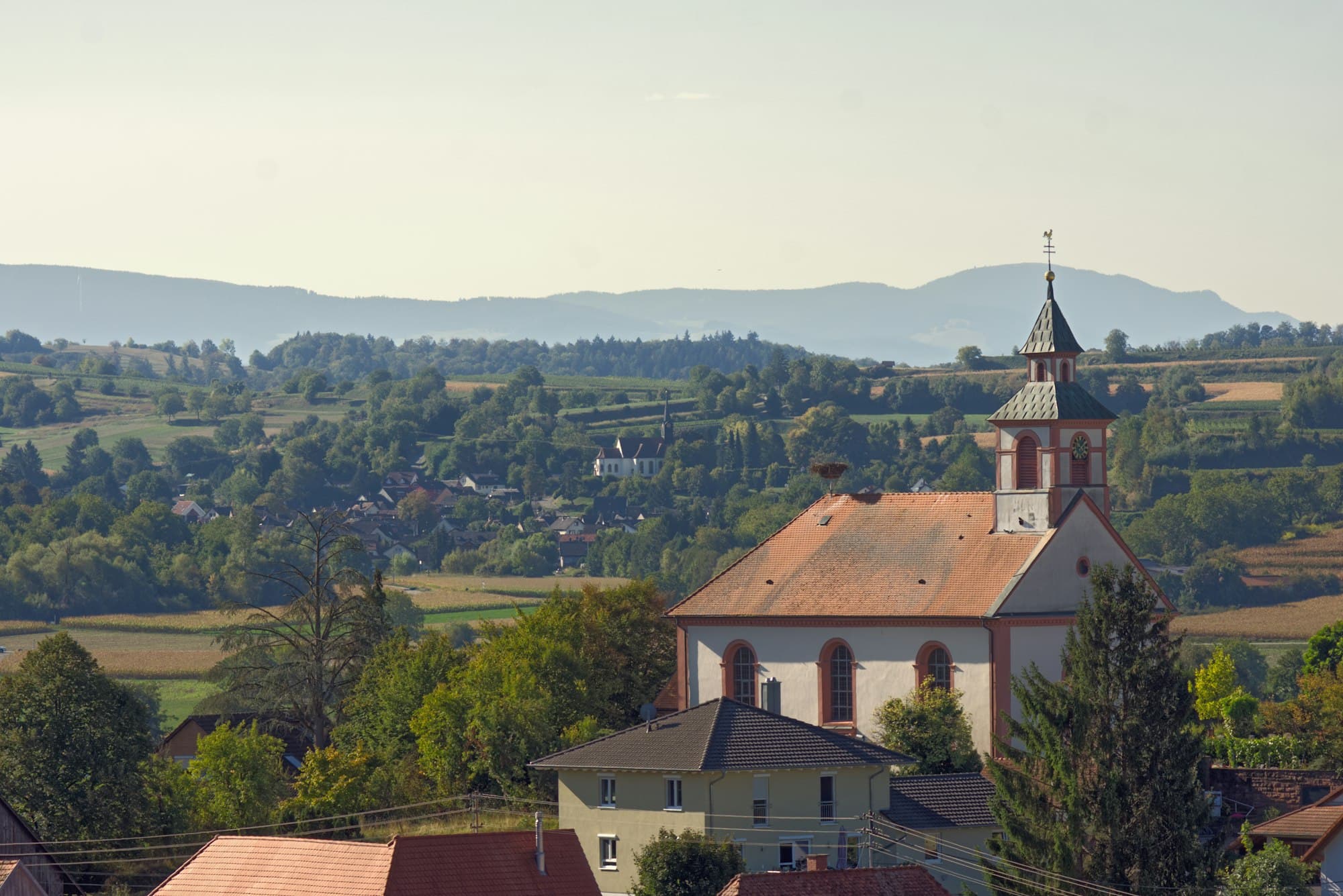 A village church with a red roof and steeple overlooking a rural valley