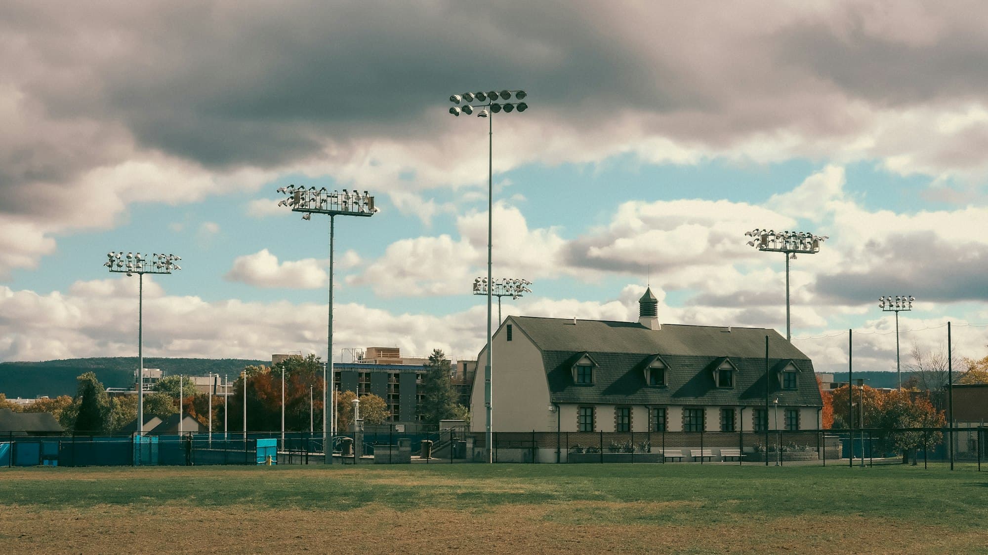 A campus sports field with tall stadium lights and a brick building under a cloudy sky