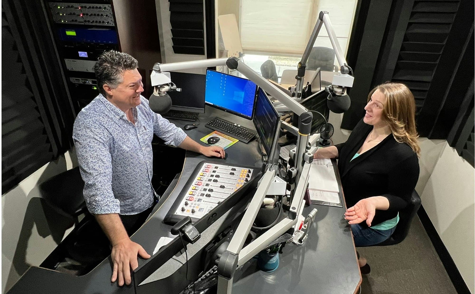 A man and a woman chatting in a radio studio with two microphones on boom arms