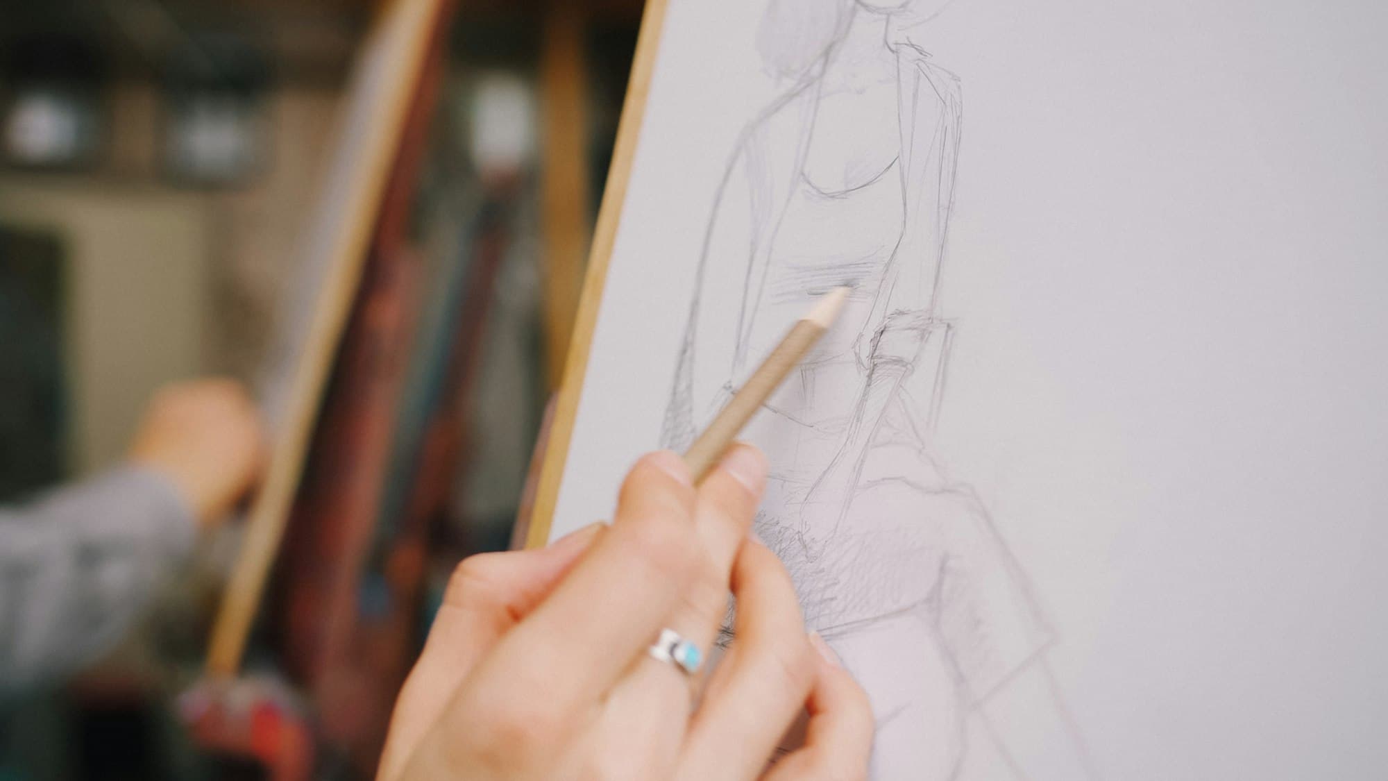 A hand holding a pencil sketching a figure on paper in an art studio