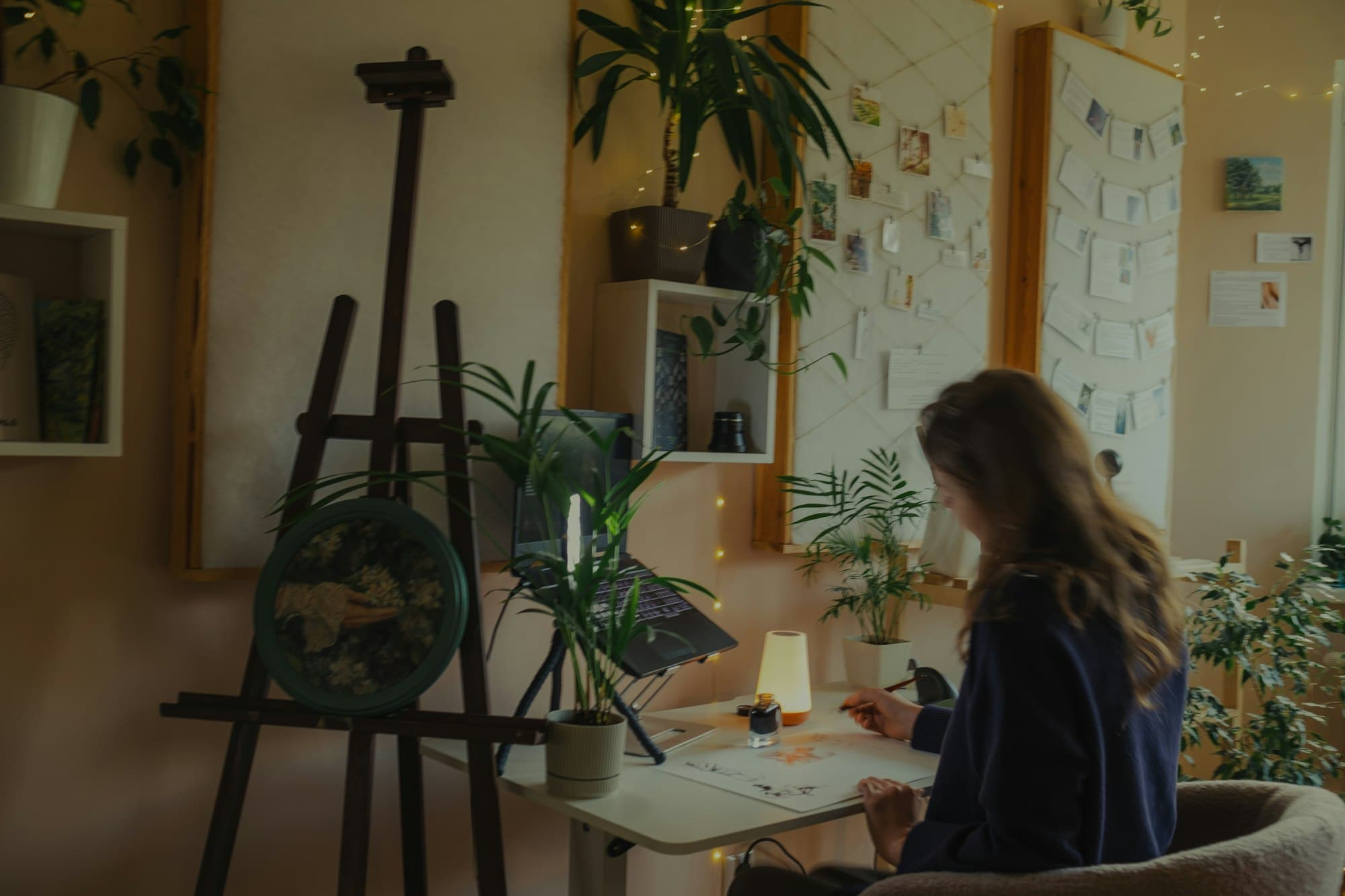 A woman drawing at a desk beside an easel in a warm, plant-filled room