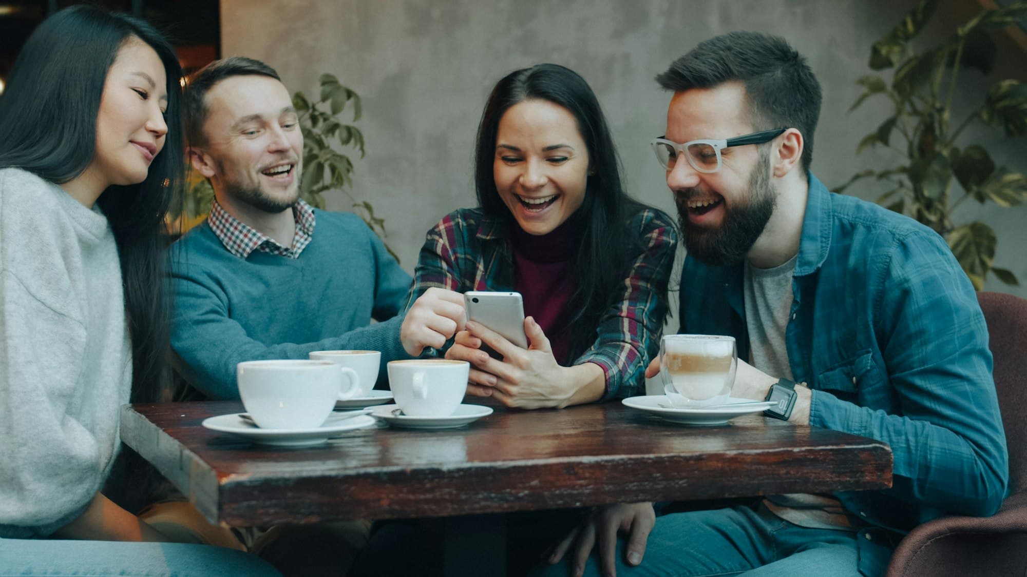Four friends laughing together at a cafe table while looking at a phone
