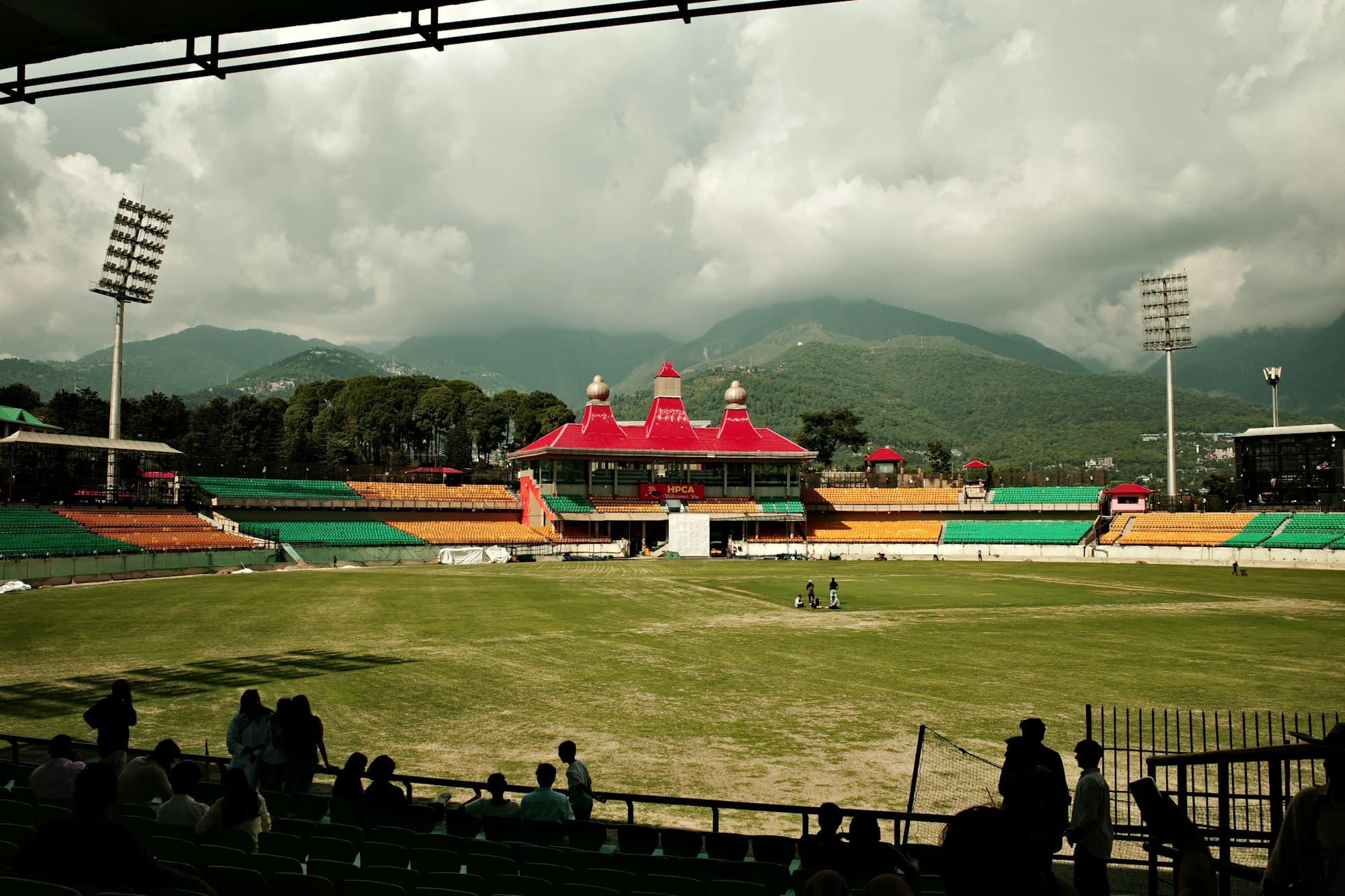 A cricket ground with green and yellow stands in front of mountains under a cloudy sky