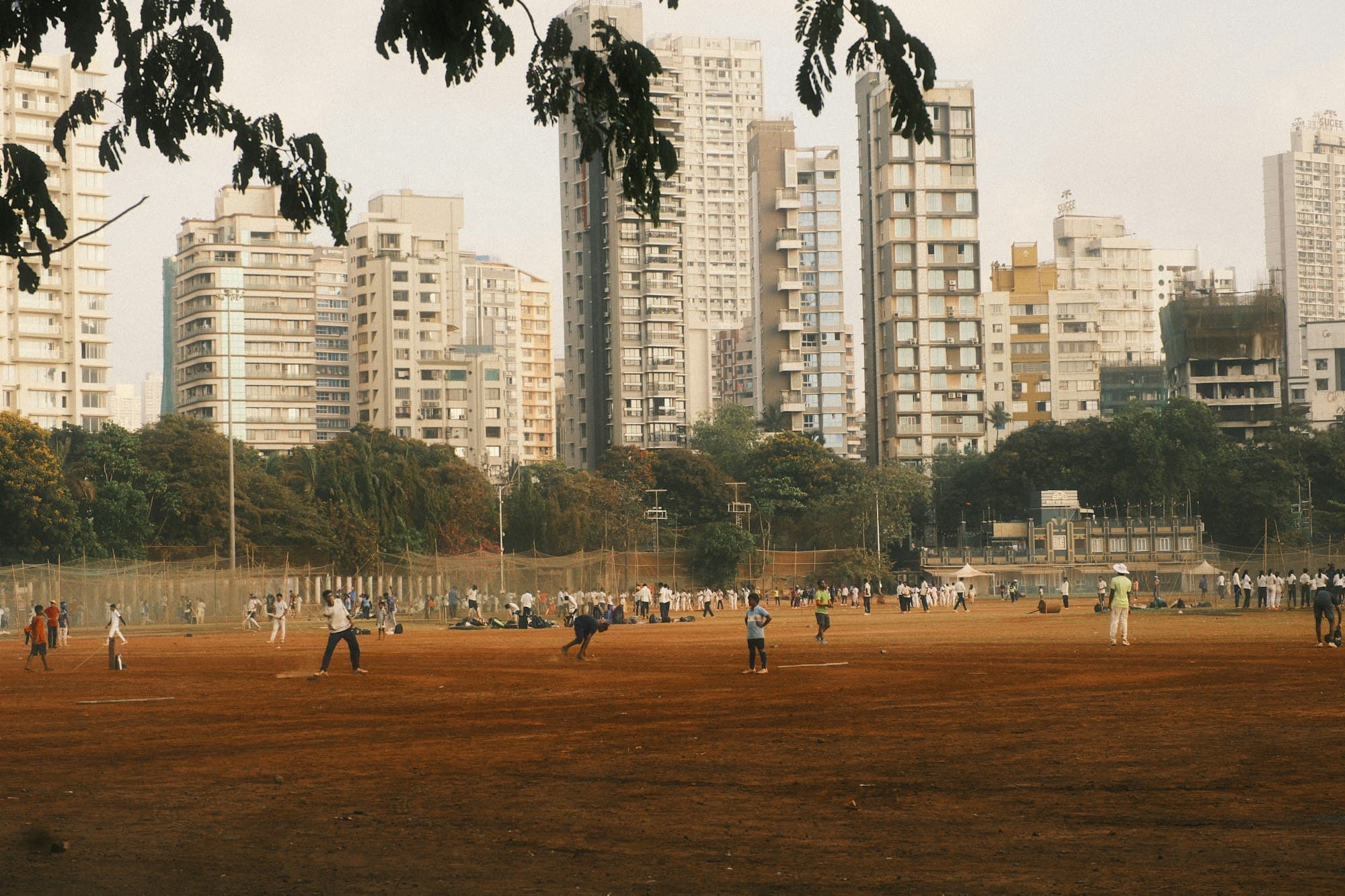 People playing cricket on a large dirt field in front of city skyscrapers