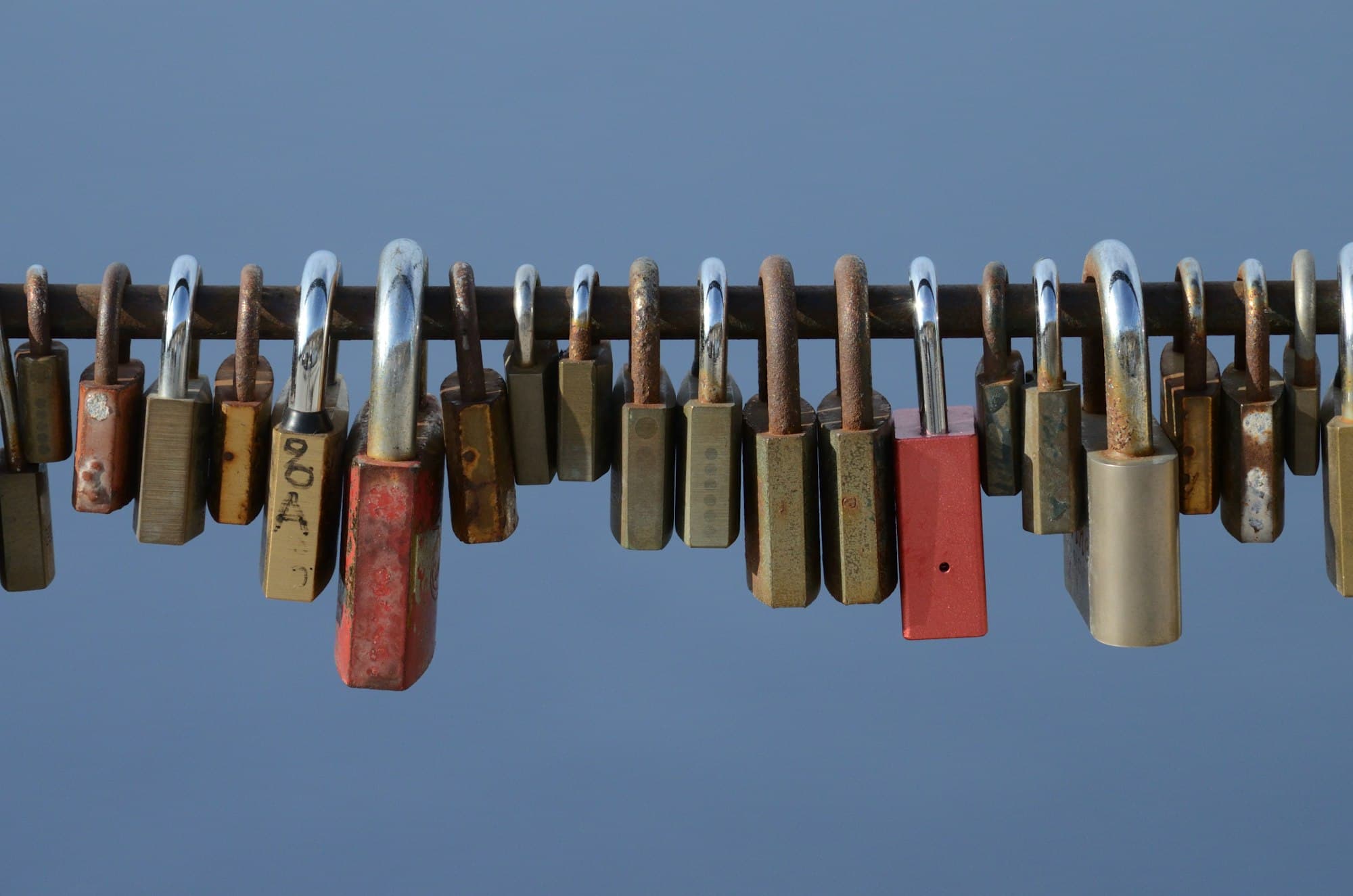 A row of rusty and brass padlocks hanging from a horizontal iron bar against a blue sky