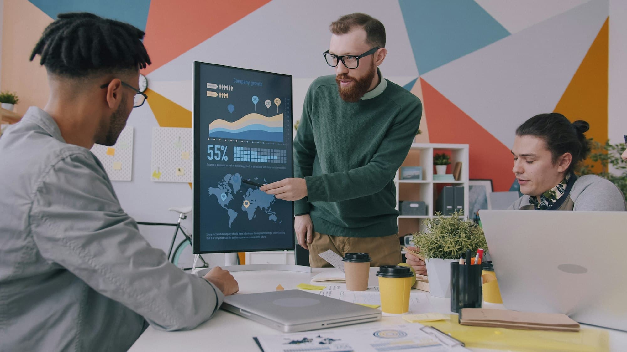 A man points at a vertical monitor showing data charts and a world map while two colleagues look on