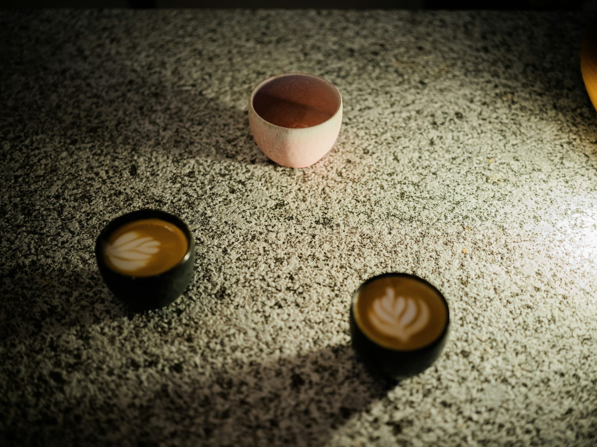 Small cups of latte art coffee and a pink ceramic cup on a speckled granite surface