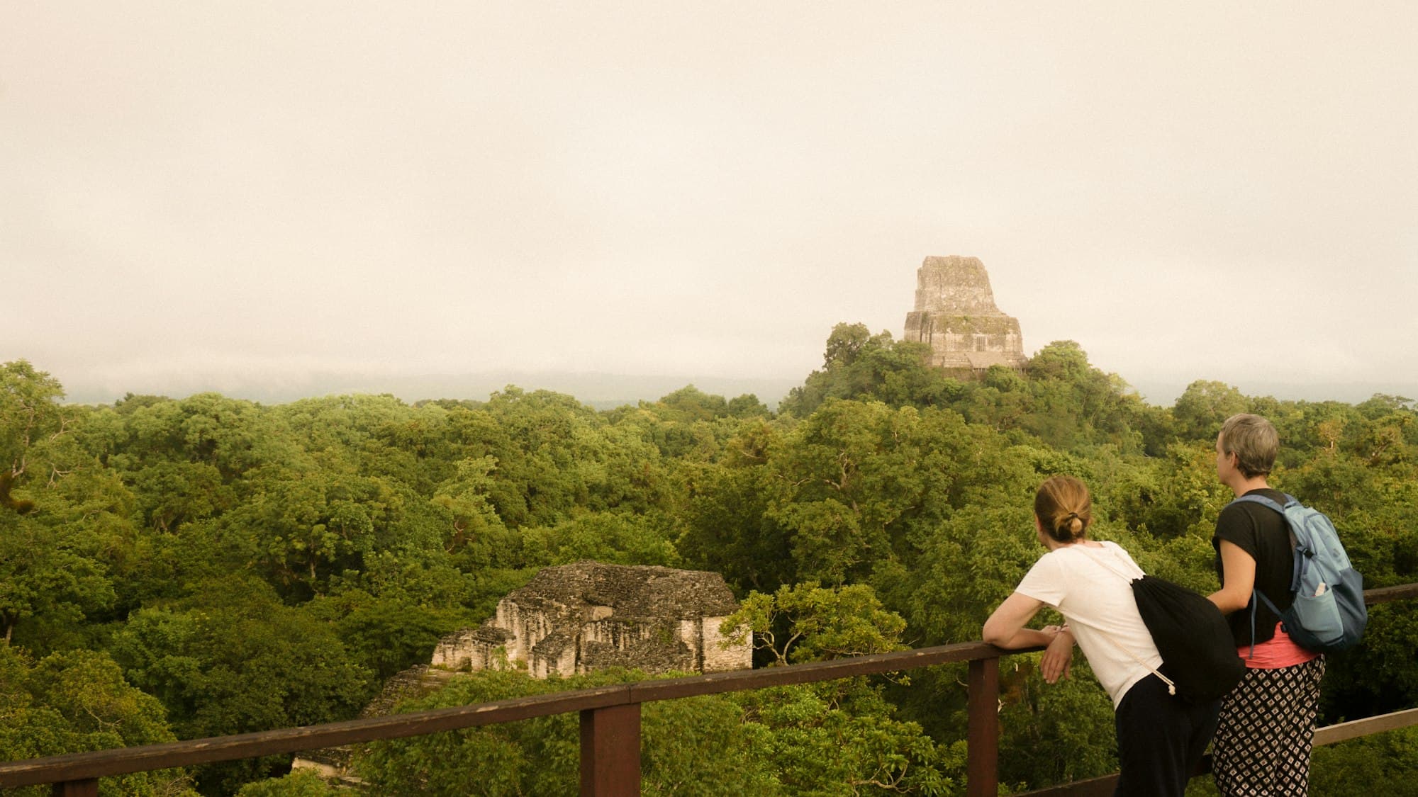 Two travelers leaning on a railing looking at ancient stone ruins above a jungle canopy