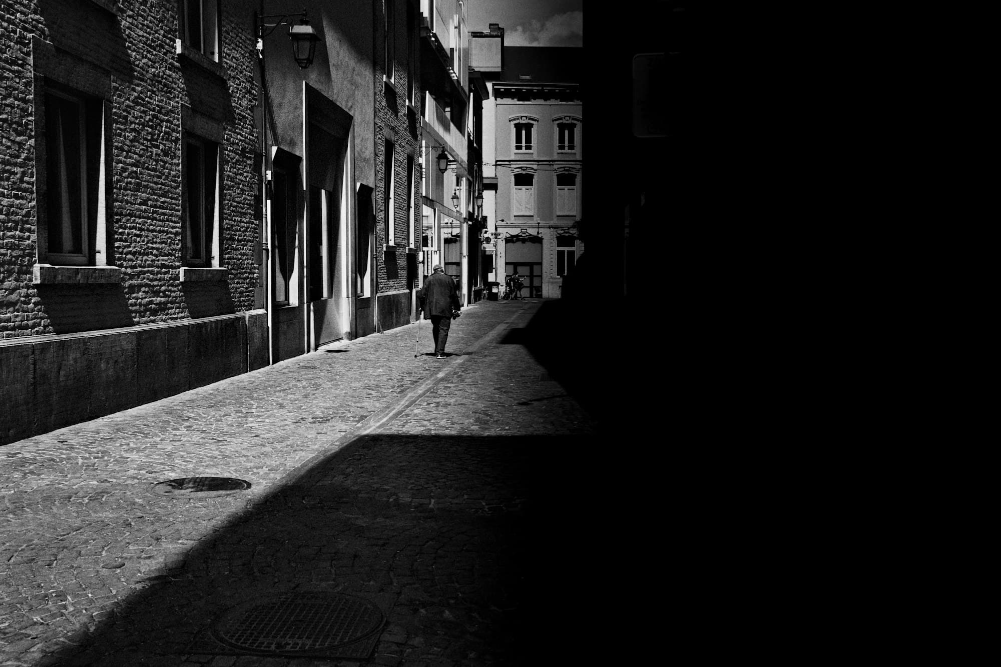 A lone figure walking down a narrow cobblestone alley in stark black-and-white light