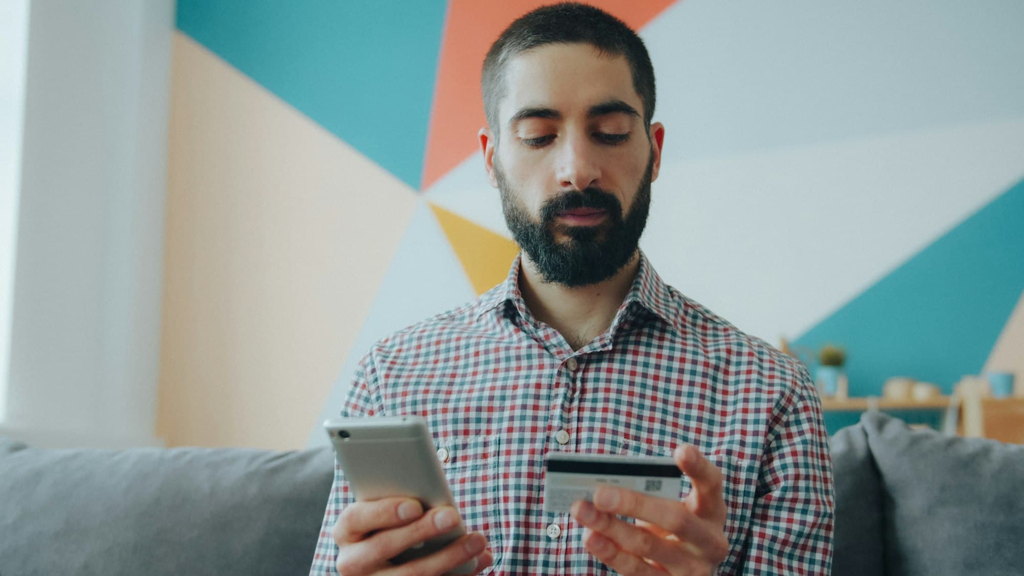 A man looking at a smartphone while holding a payment card on a sofa
