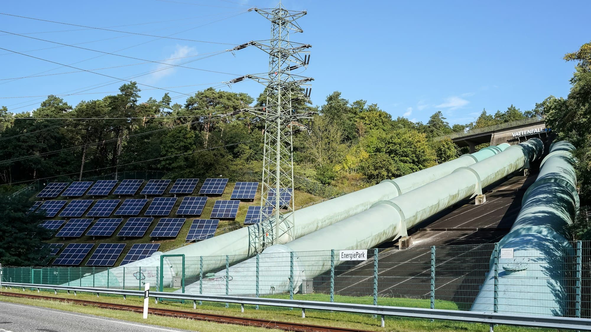 Large green pipes and solar panels on a hillside beside a power pylon