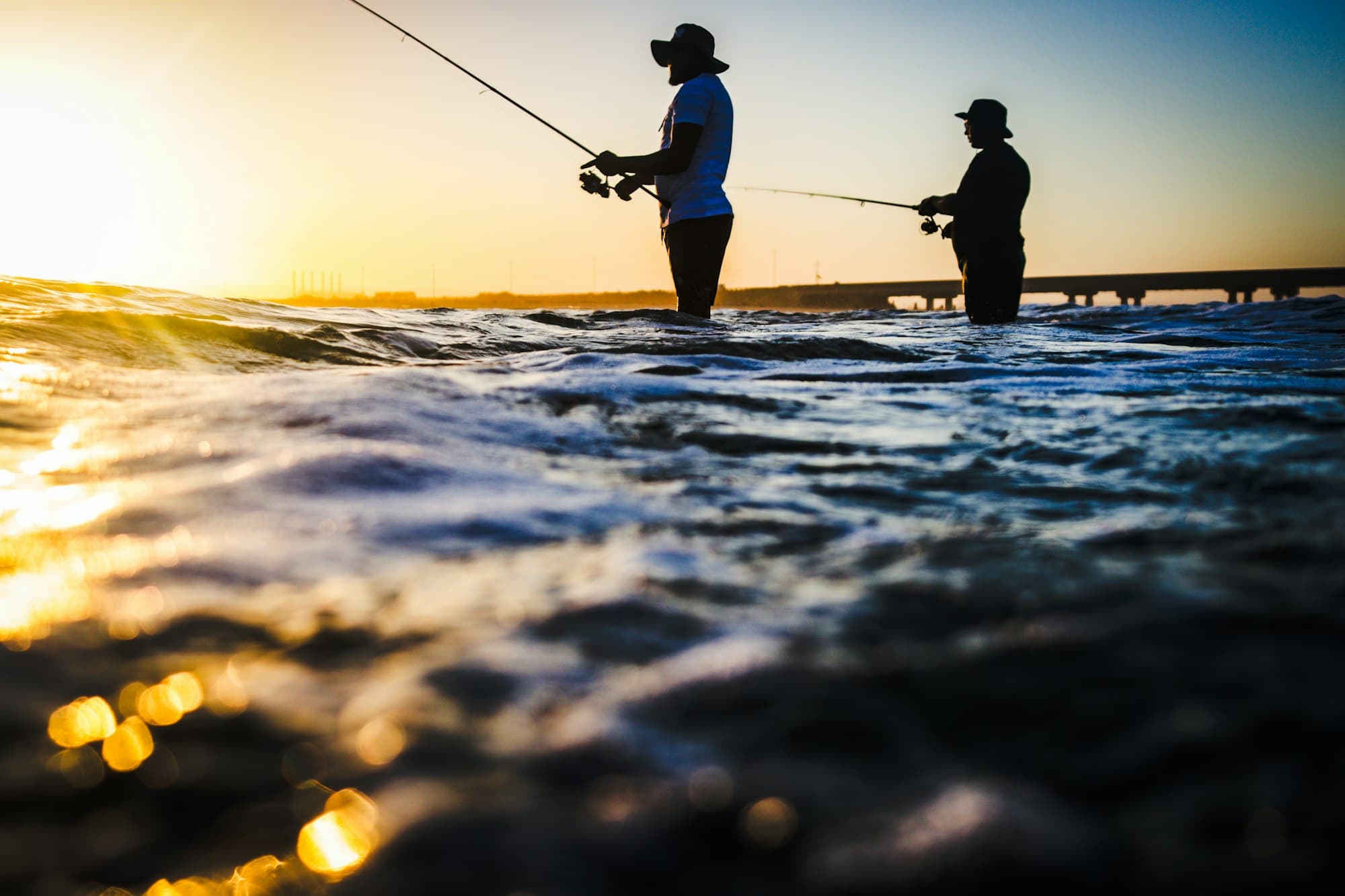 Two anglers in silhouette casting lines into the ocean at golden hour
