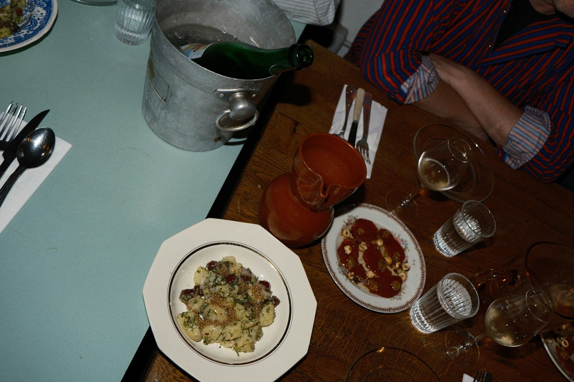 A dinner table with potato salad, sliced meat, wine glasses and a bottle in an ice bucket