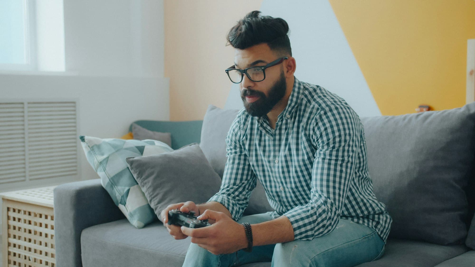 A bearded man in glasses holding a game controller on a grey sofa