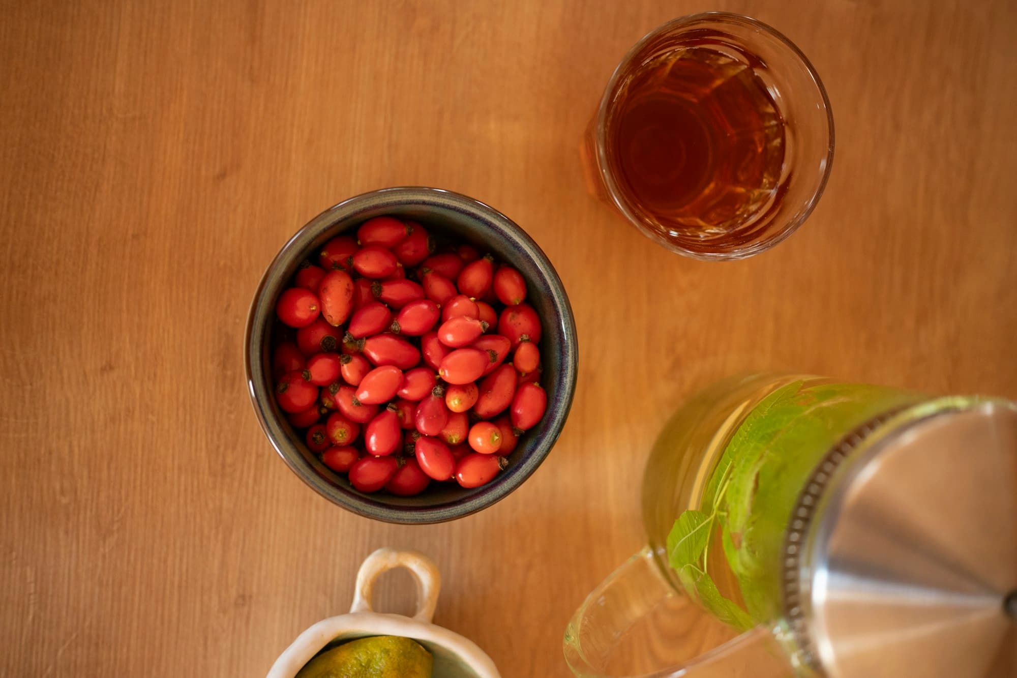 A bowl of red rose hips beside a glass of herbal tea on a wooden table