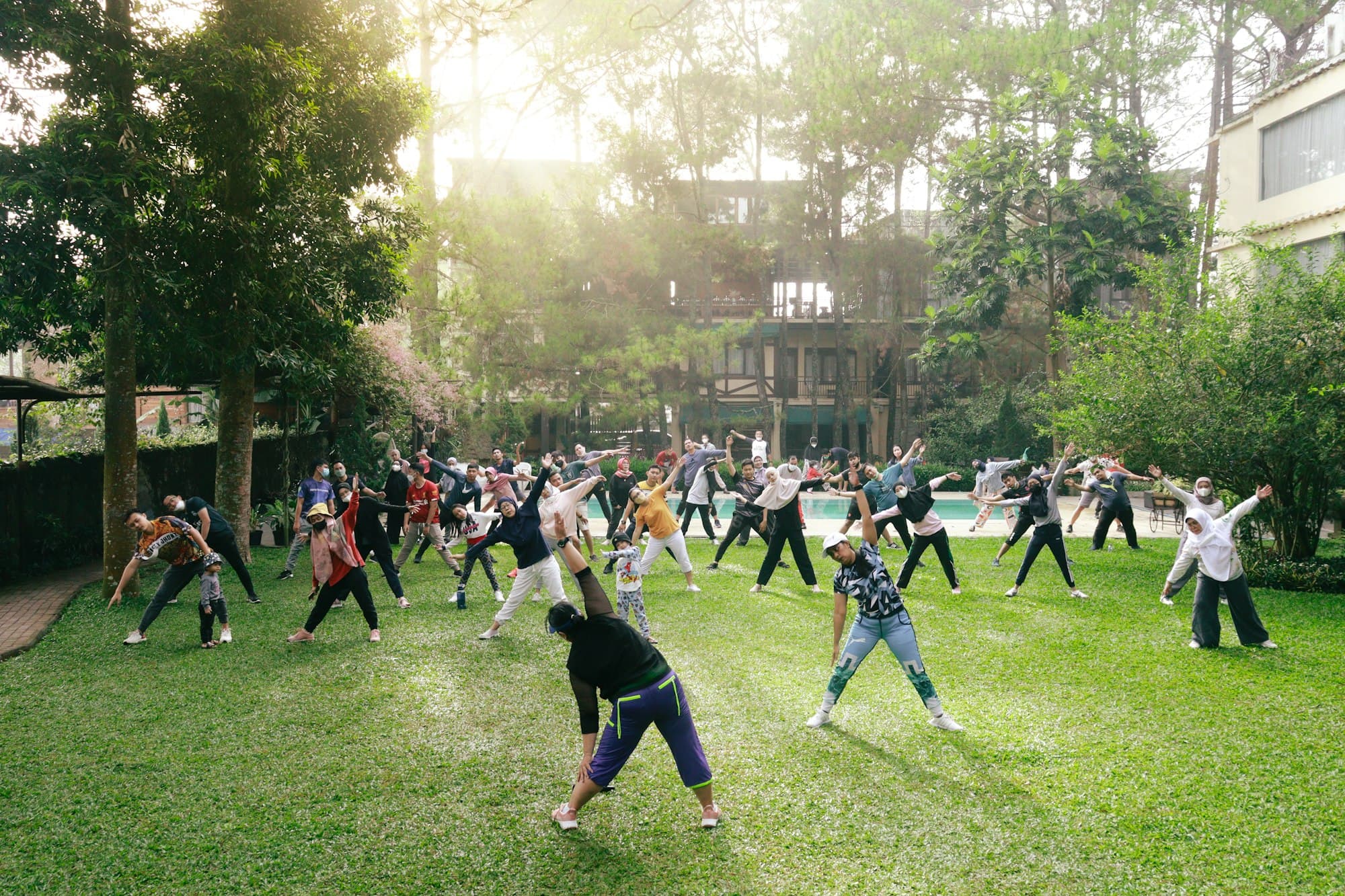 A group of people stretching together on a sunlit green lawn among trees