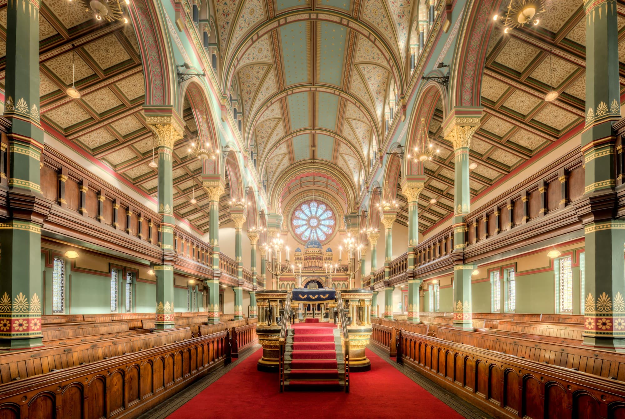 An ornate synagogue interior with a red carpet aisle, wooden pews and a round stained-glass window