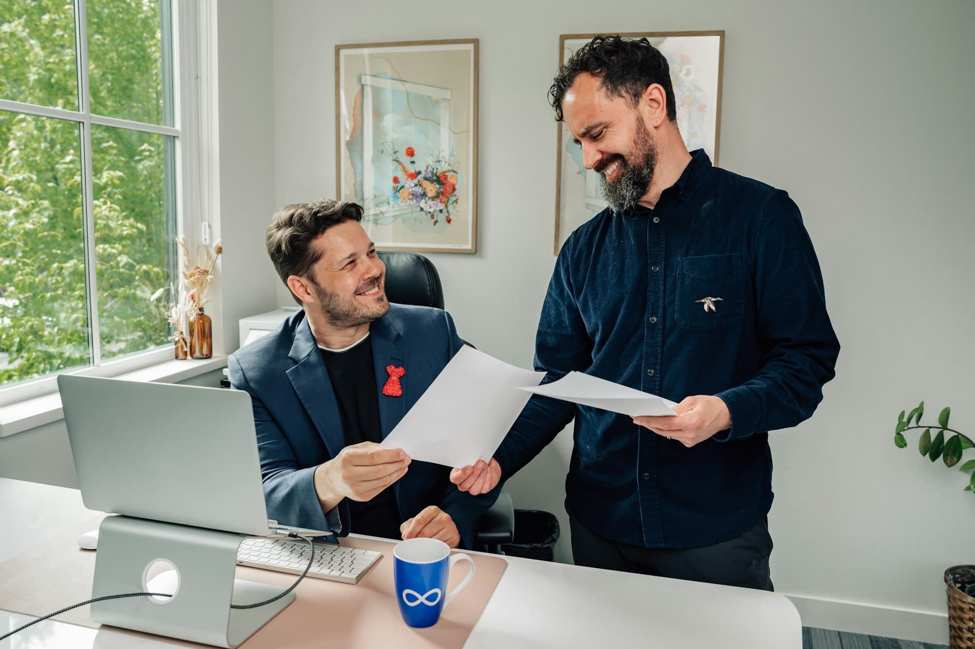 A standing colleague talks through papers with a seated colleague at a bright office desk