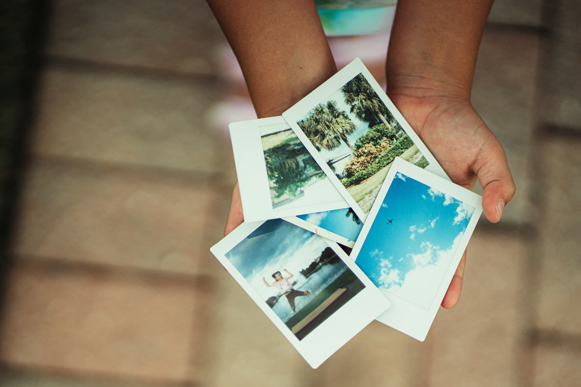 Two hands hold out a fanned stack of instant photographs of palm trees and blue sky