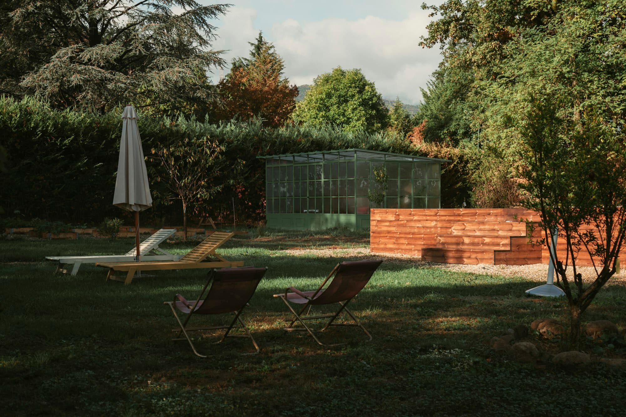 Lounge chairs and a closed parasol on a shady garden lawn beside a greenhouse