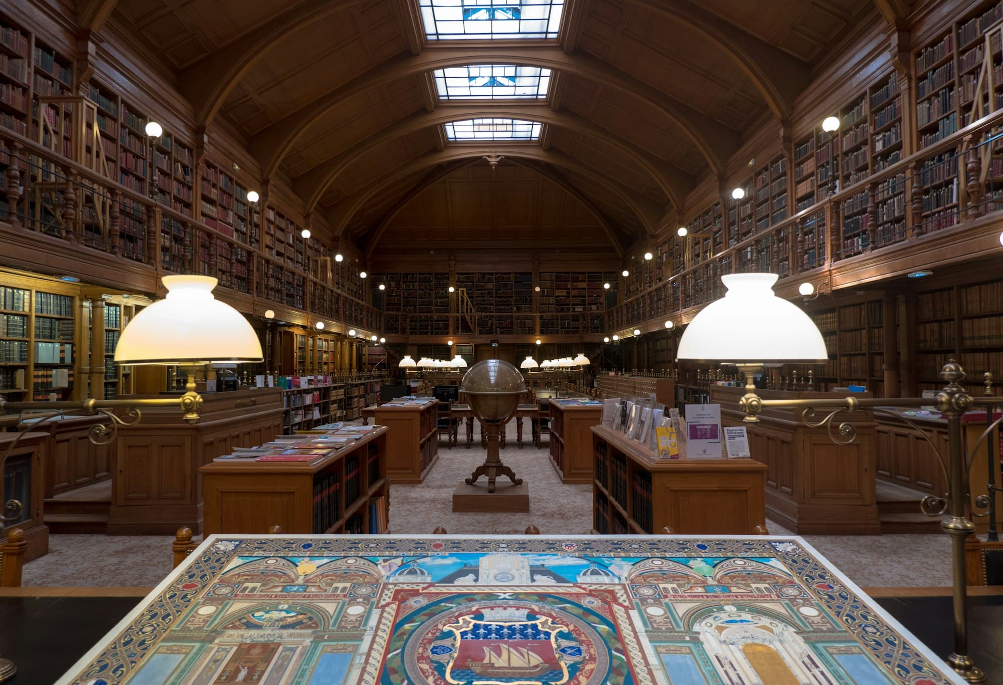 A historic library with tall wooden shelves, a central globe and glowing desk lamps