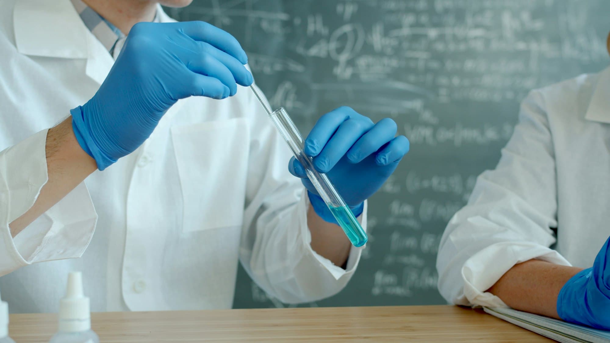 Scientists in white lab coats and blue gloves holding a test tube in front of a chalkboard of formulas