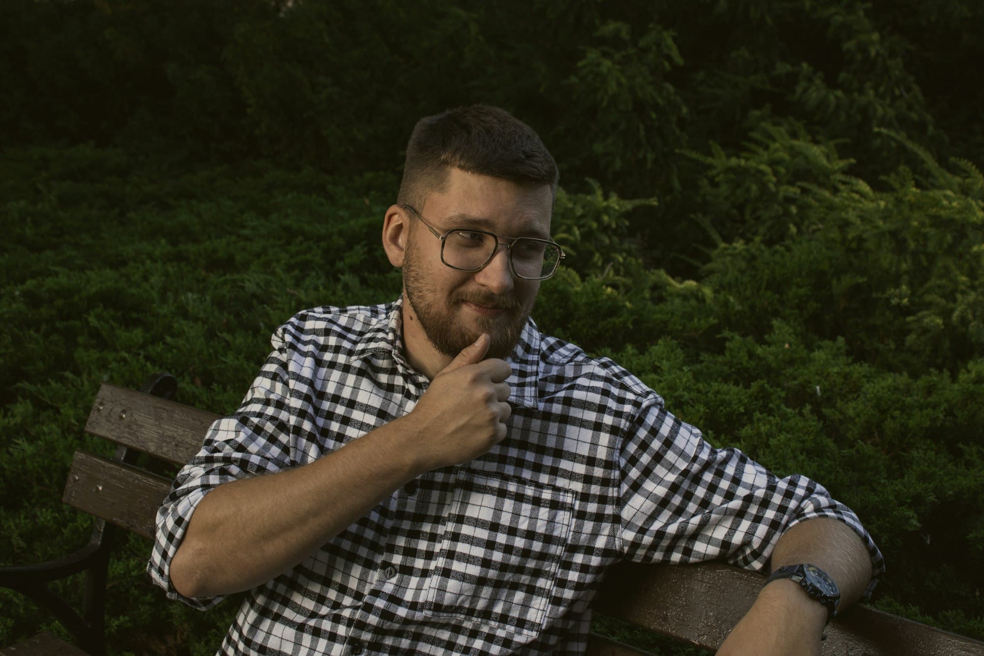 A bearded man in glasses and a plaid shirt relaxing on a park bench among greenery