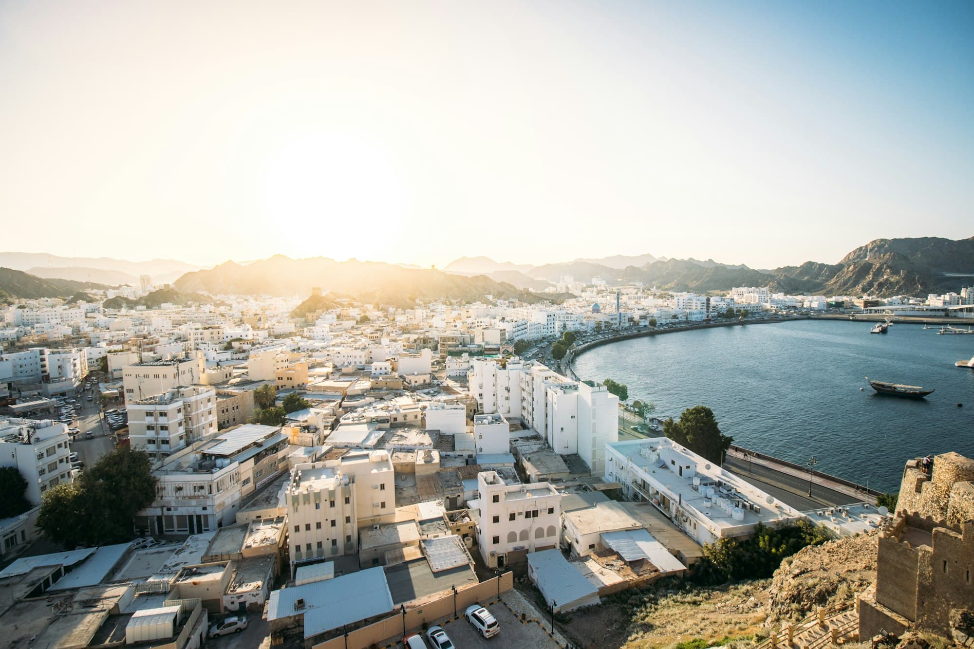 White buildings of Muscat curving around a harbour with a dhow, rugged mountains behind at sunset