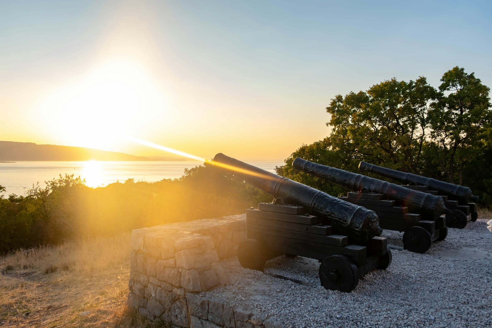 Three black cannons on stone mounts overlooking a bay at low sun