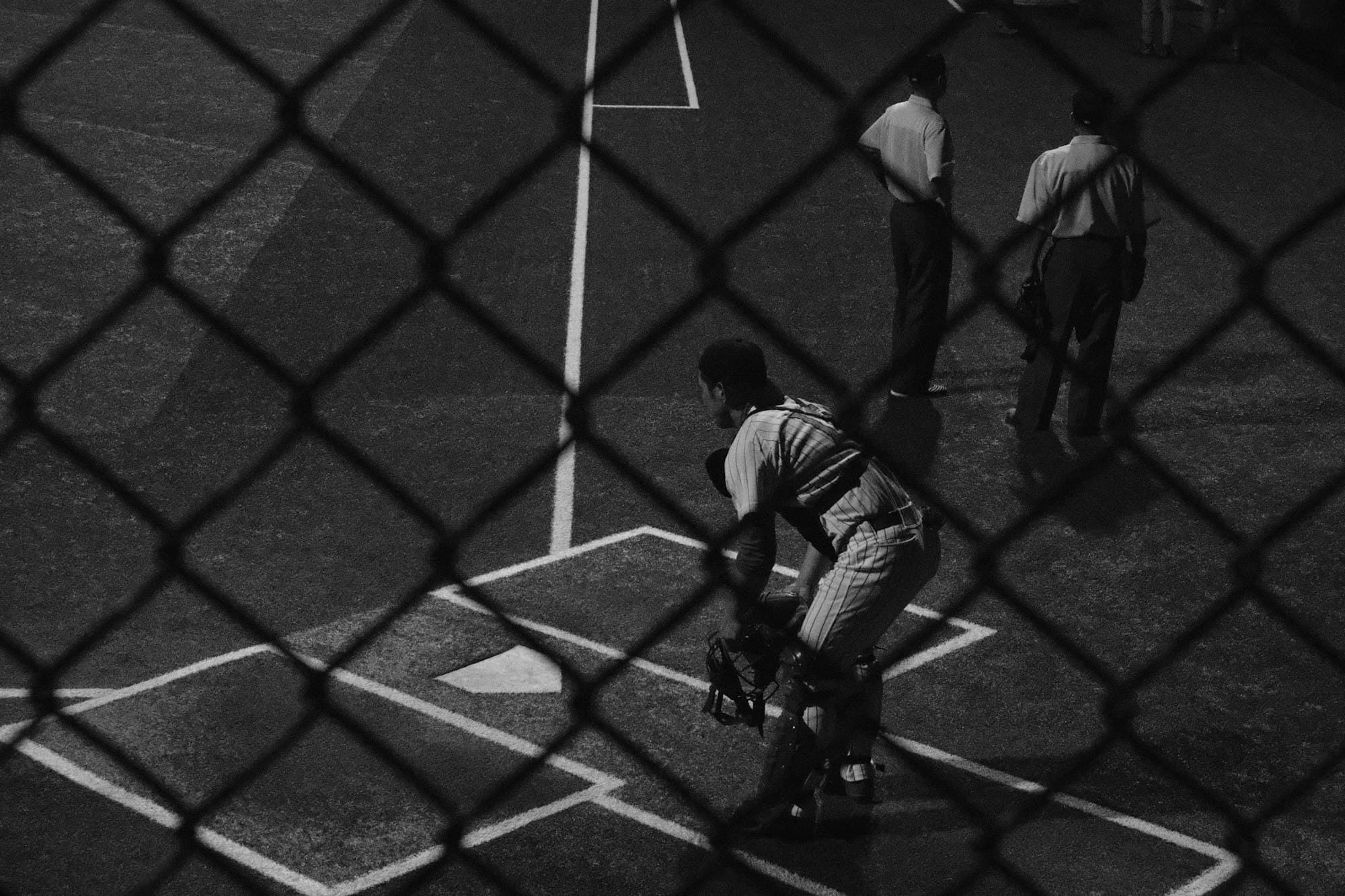 A baseball catcher crouches behind home plate seen through a chain-link fence, in black and white