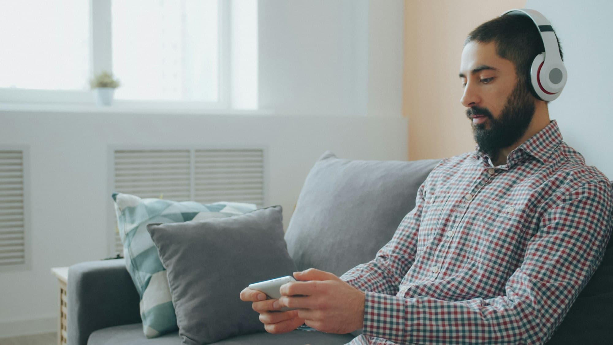 A bearded man wearing white headphones relaxing on a sofa with a phone