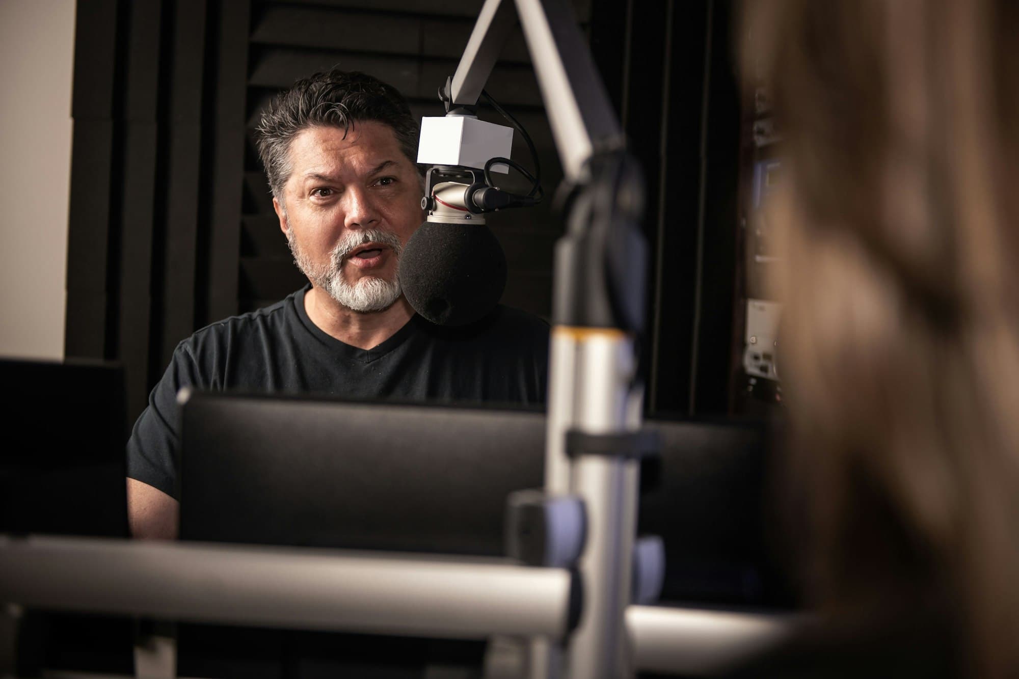 A man talking at a studio microphone on a boom arm in a sound-treated room