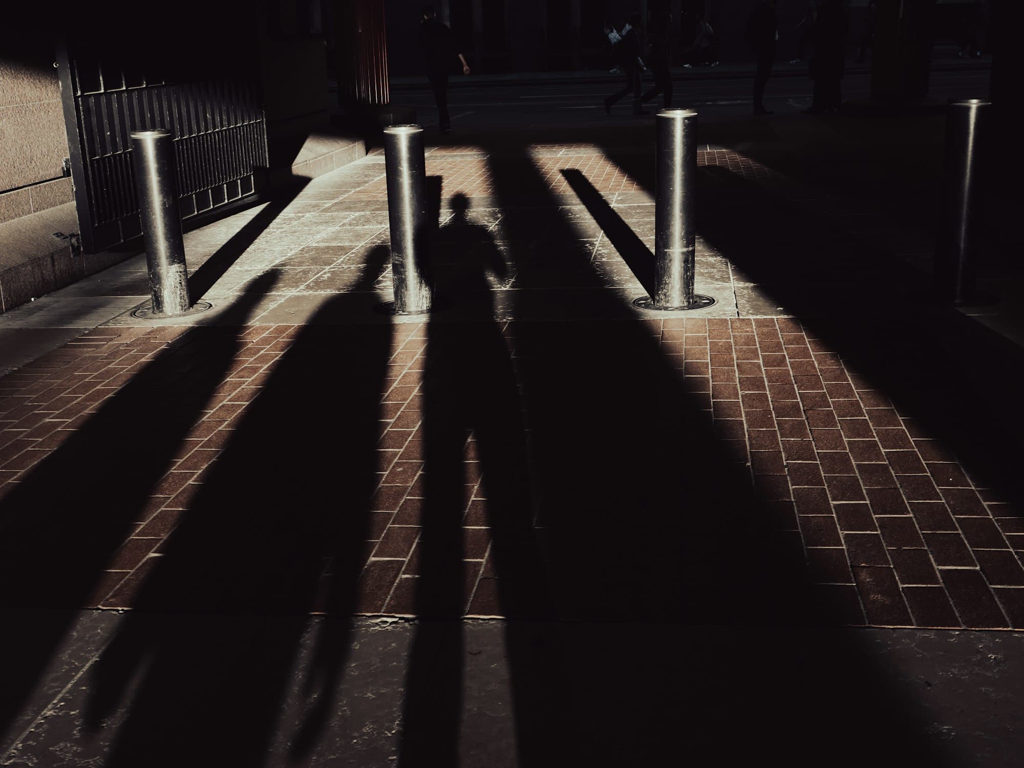 Long dark shadows of people cast across a brick walkway with metal bollards