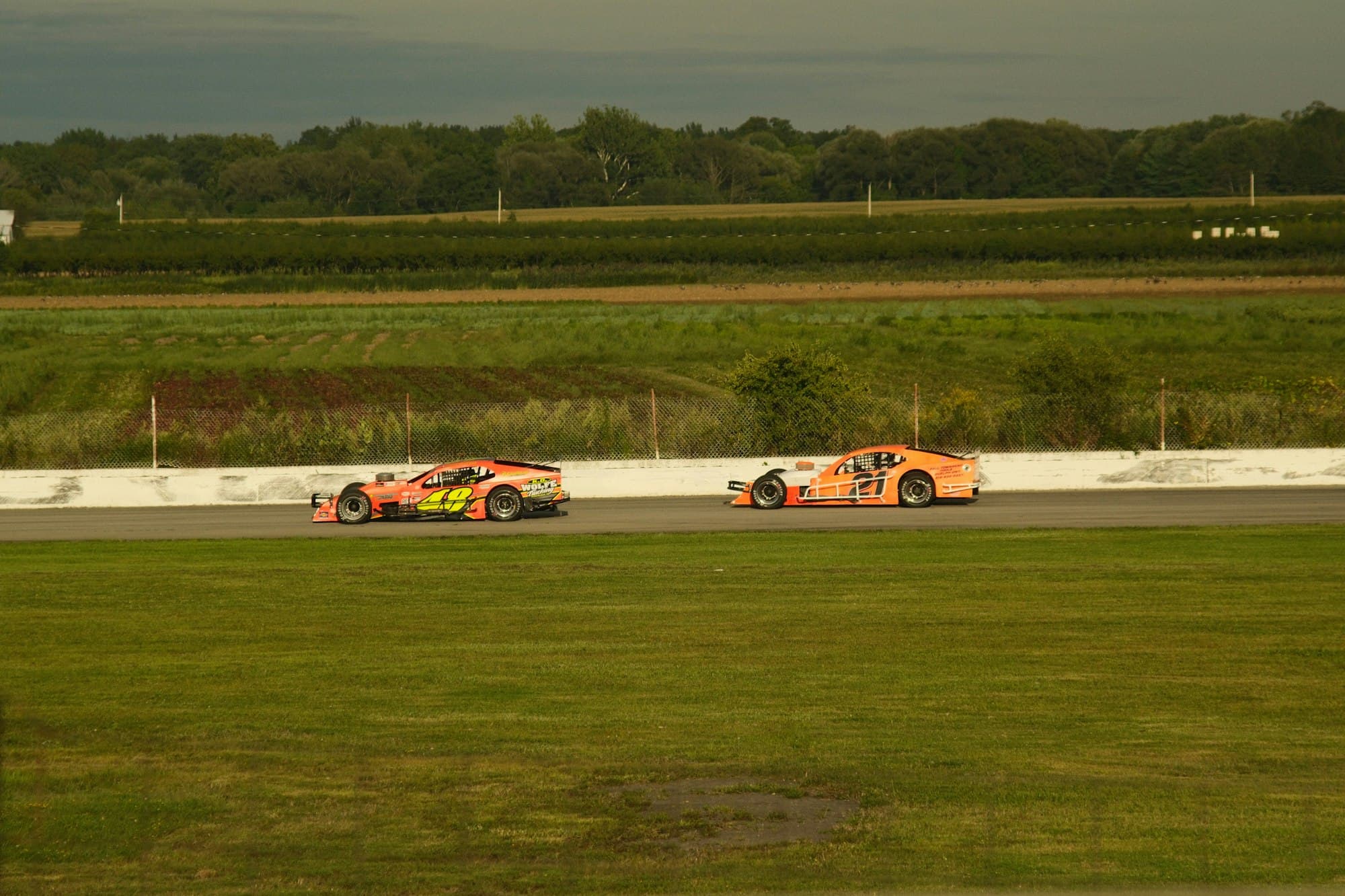 Two orange stock cars racing on a paved oval beside a green field