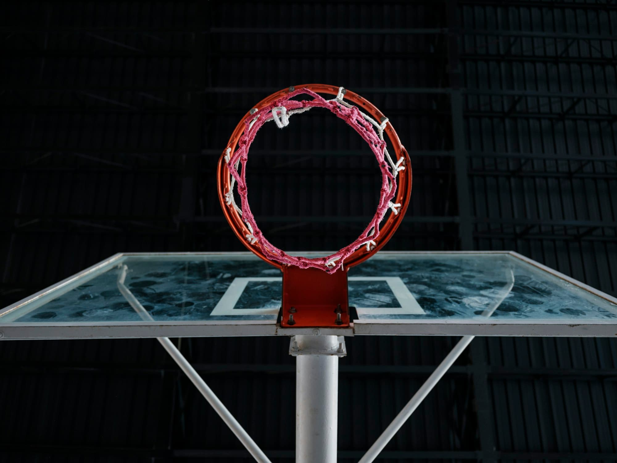A basketball rim with a pink net on a glass backboard in a dark indoor gym