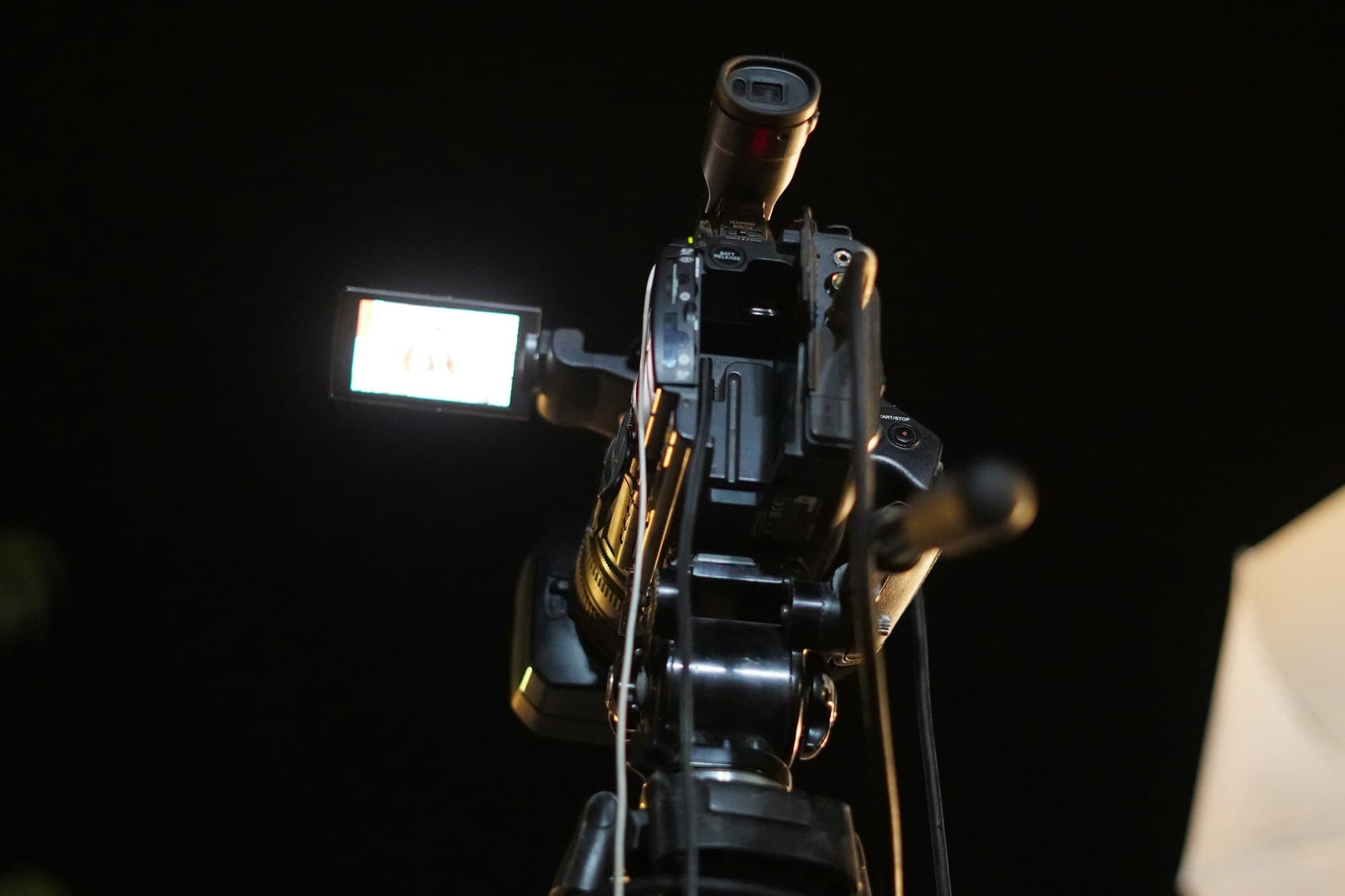 A professional video camera on a tripod with its screen lit against black