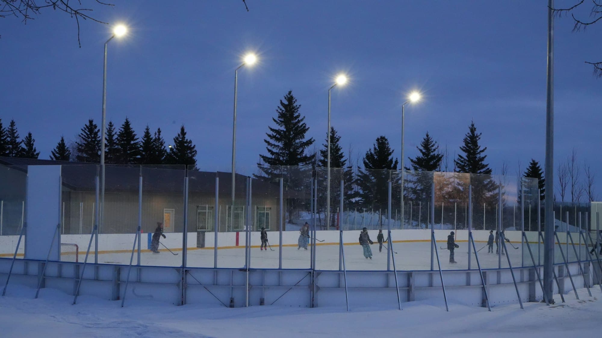 An outdoor ice hockey rink lit by floodlights at dusk with distant skaters