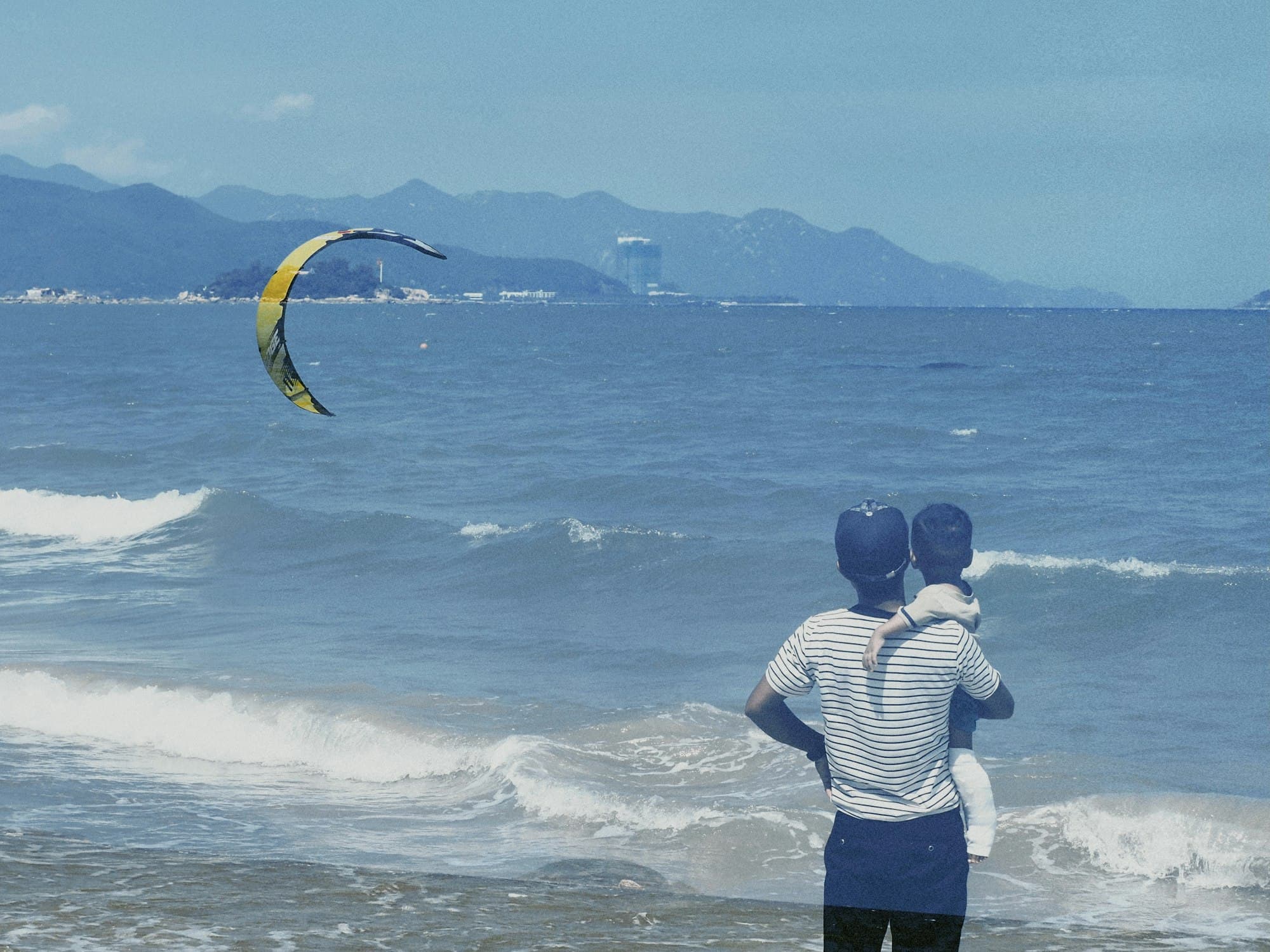 A parent seen from behind holds a toddler at the water's edge watching a kite surfer