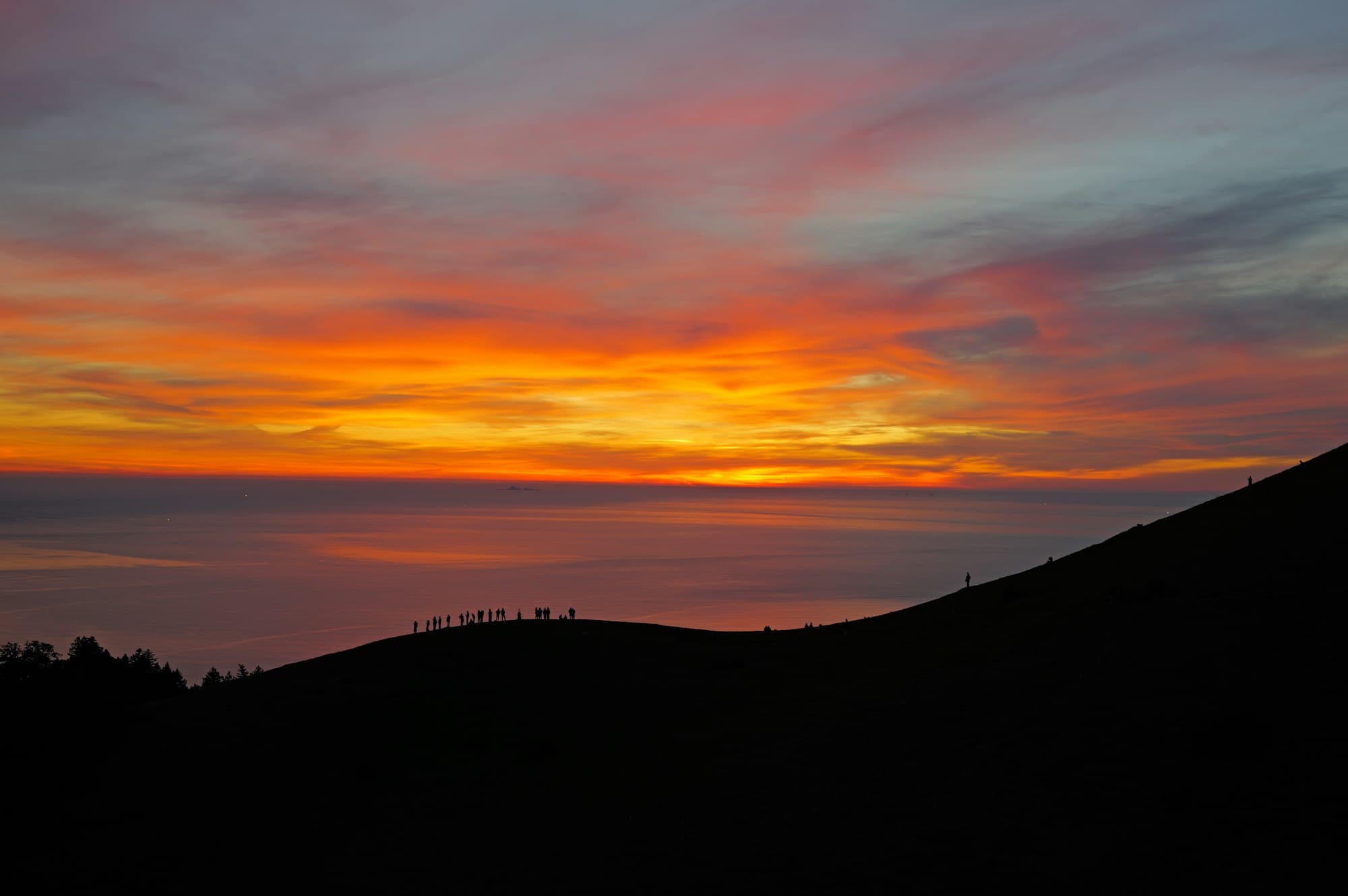 Small silhouettes of people on a hilltop ridge against a vivid orange sky over the sea