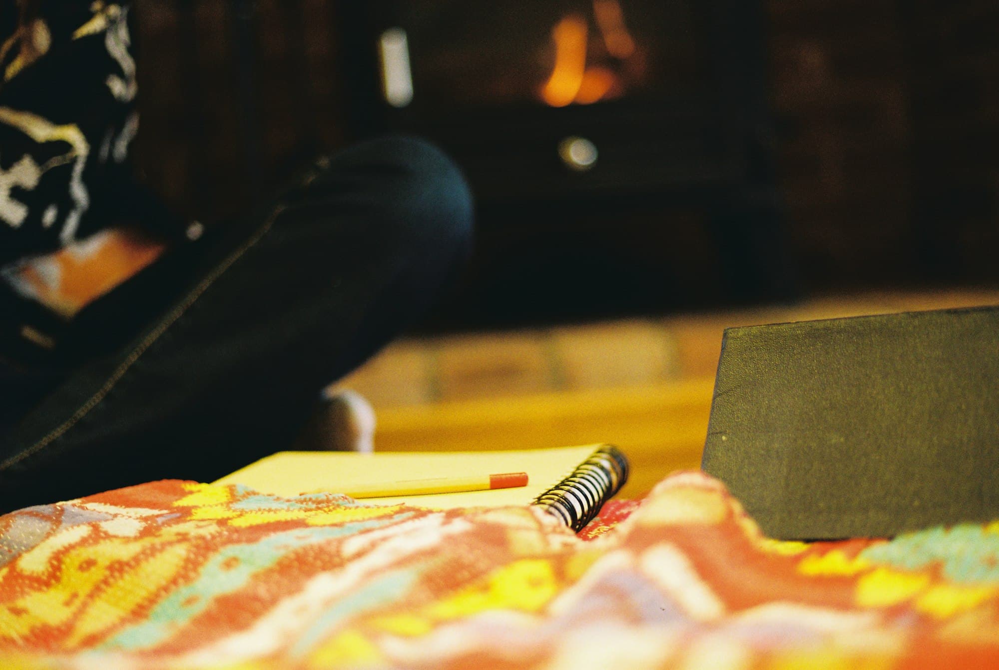 An open notebook and pencil on a colourful patterned throw in front of a softly glowing fire