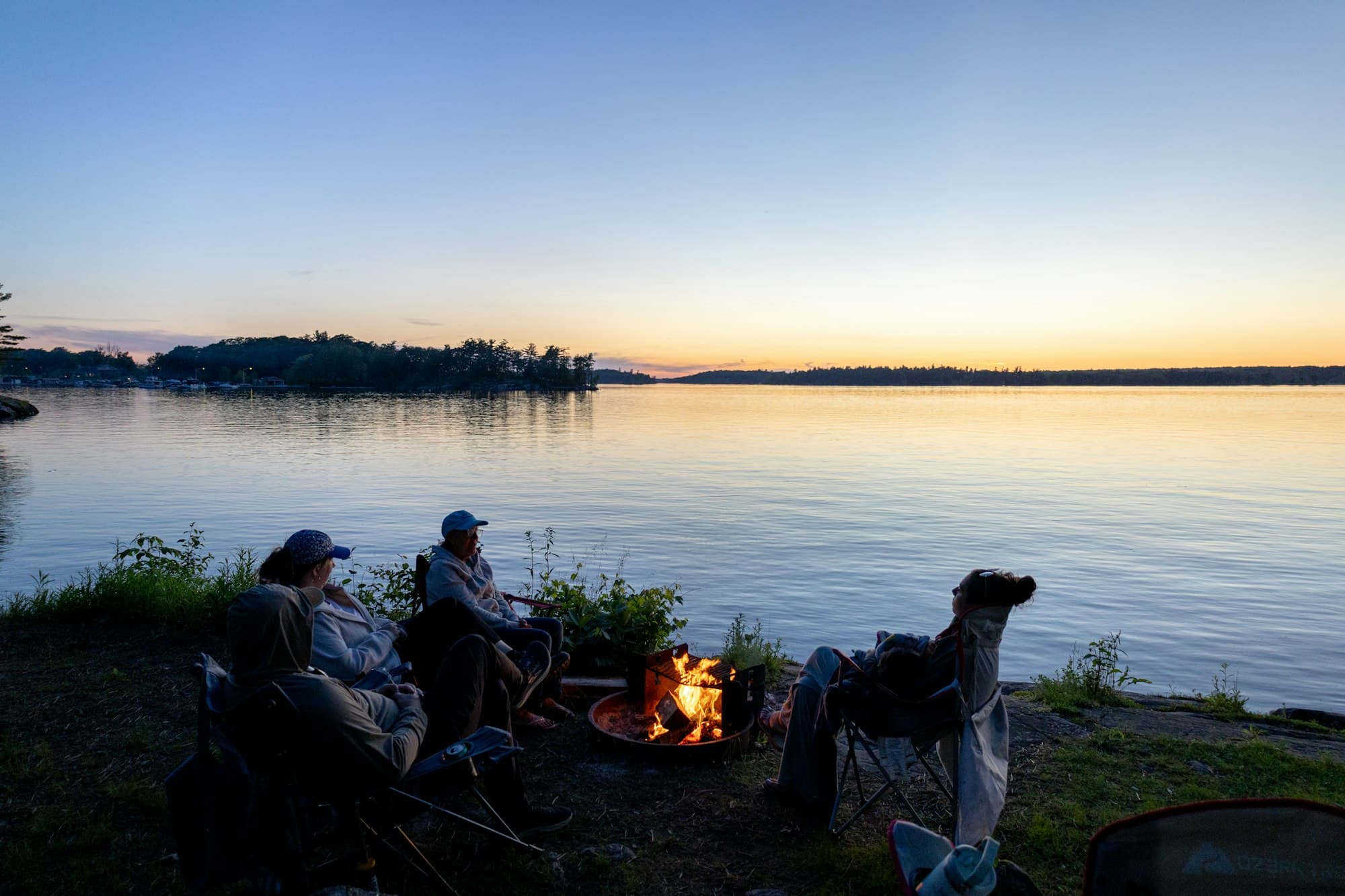 People sitting in camp chairs around a campfire beside a lake at sunset