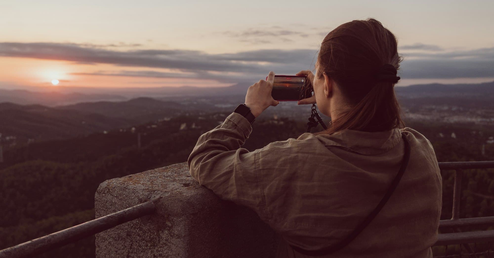 A person seen from behind photographing a low sun over rolling hills