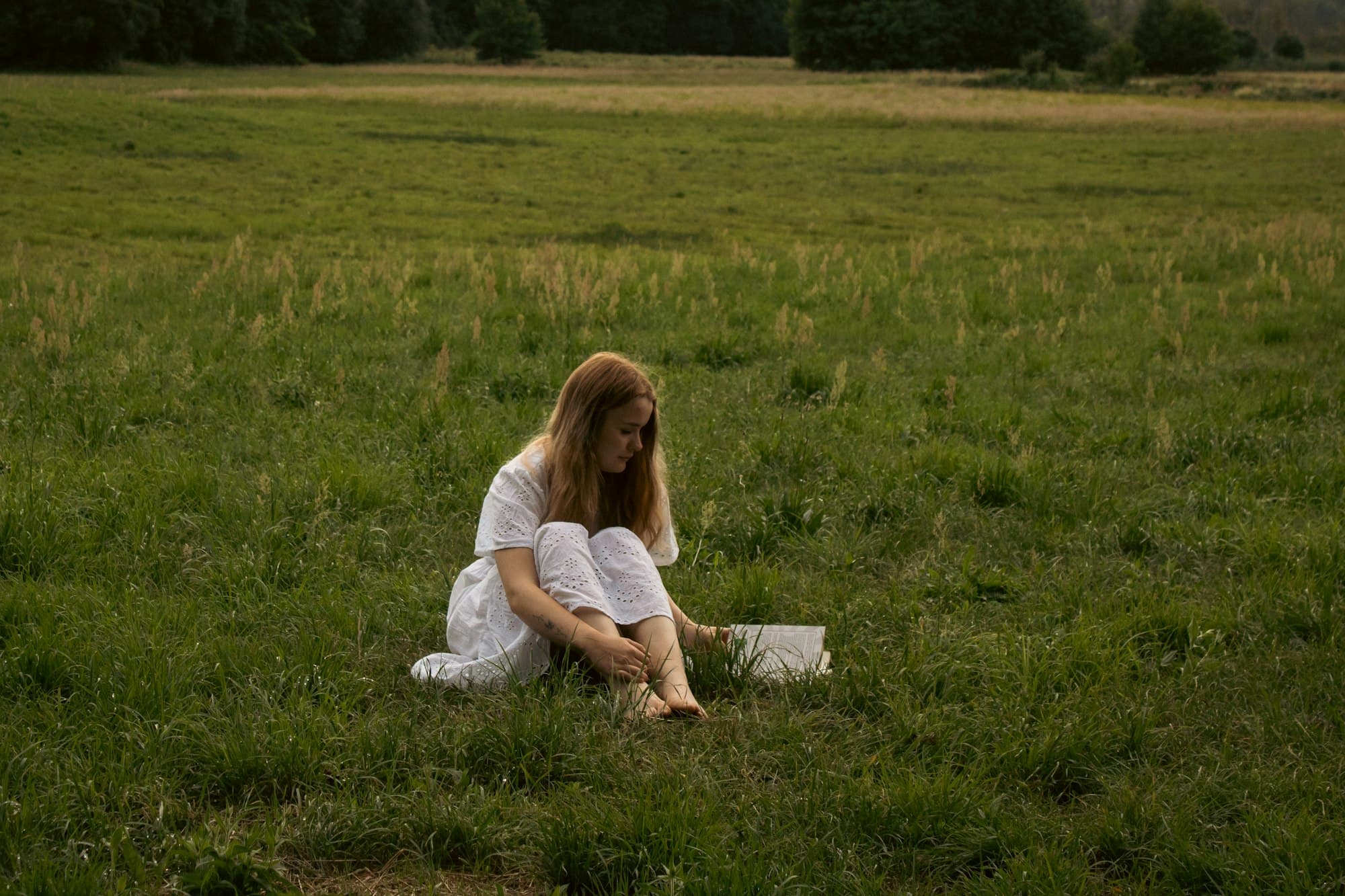 A woman in a white dress sits reading a small book in a wide green meadow