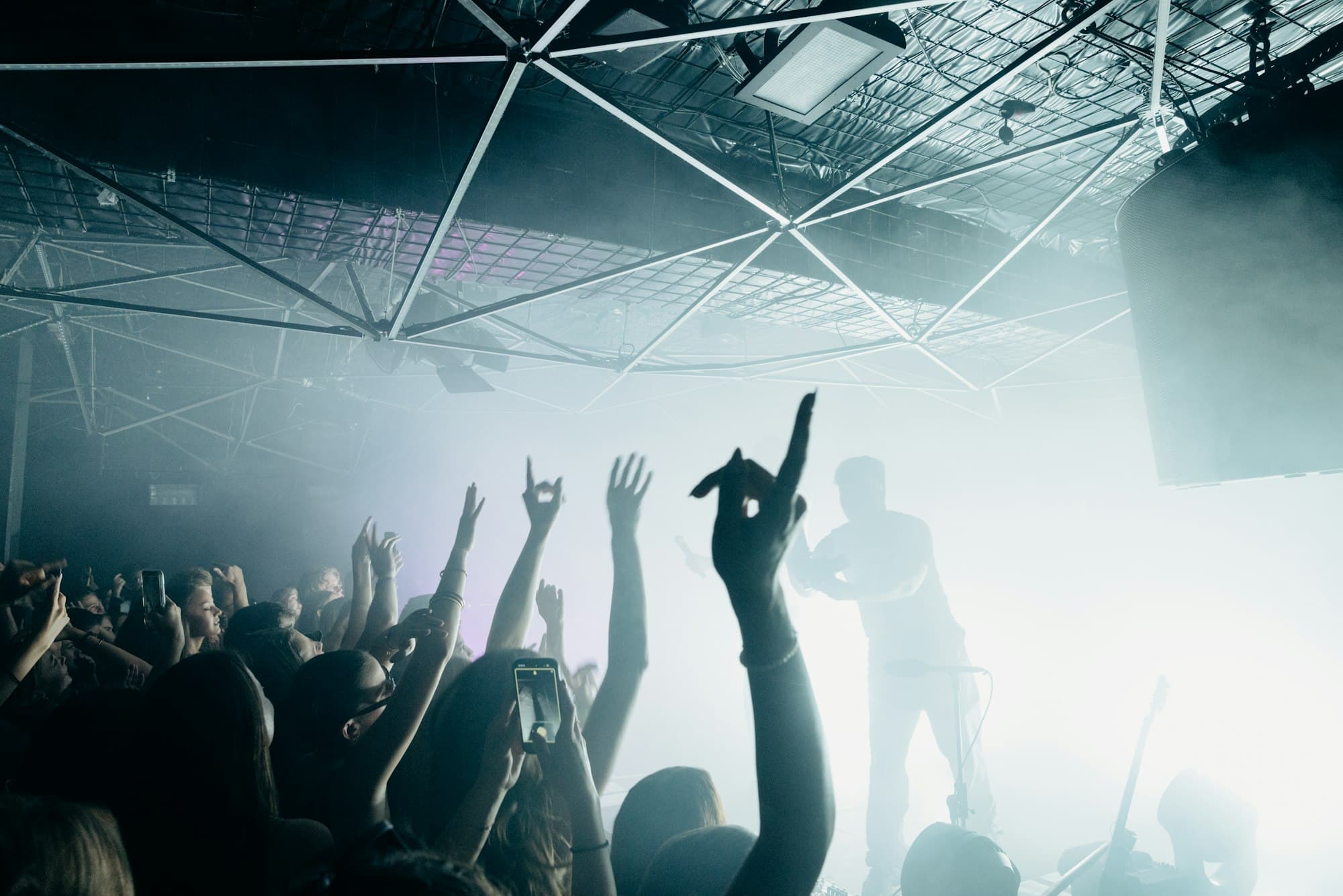 A crowd with raised hands and phones at a live concert in a hazy, bright-lit venue