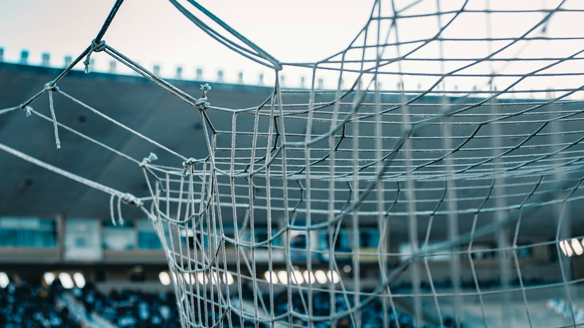 A white soccer goal net in the foreground with a blurred stadium behind