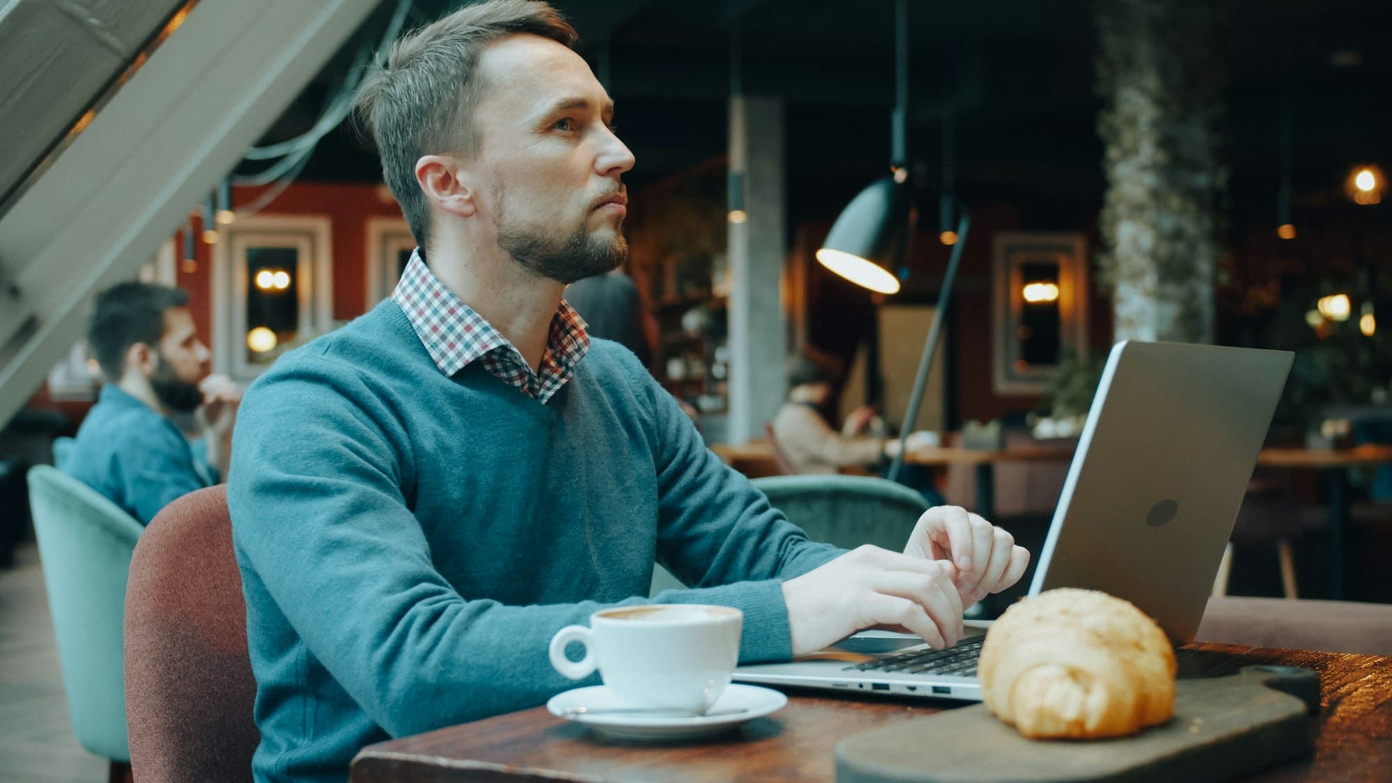 A man in a blue sweater working on a laptop in a cafe with coffee and a croissant