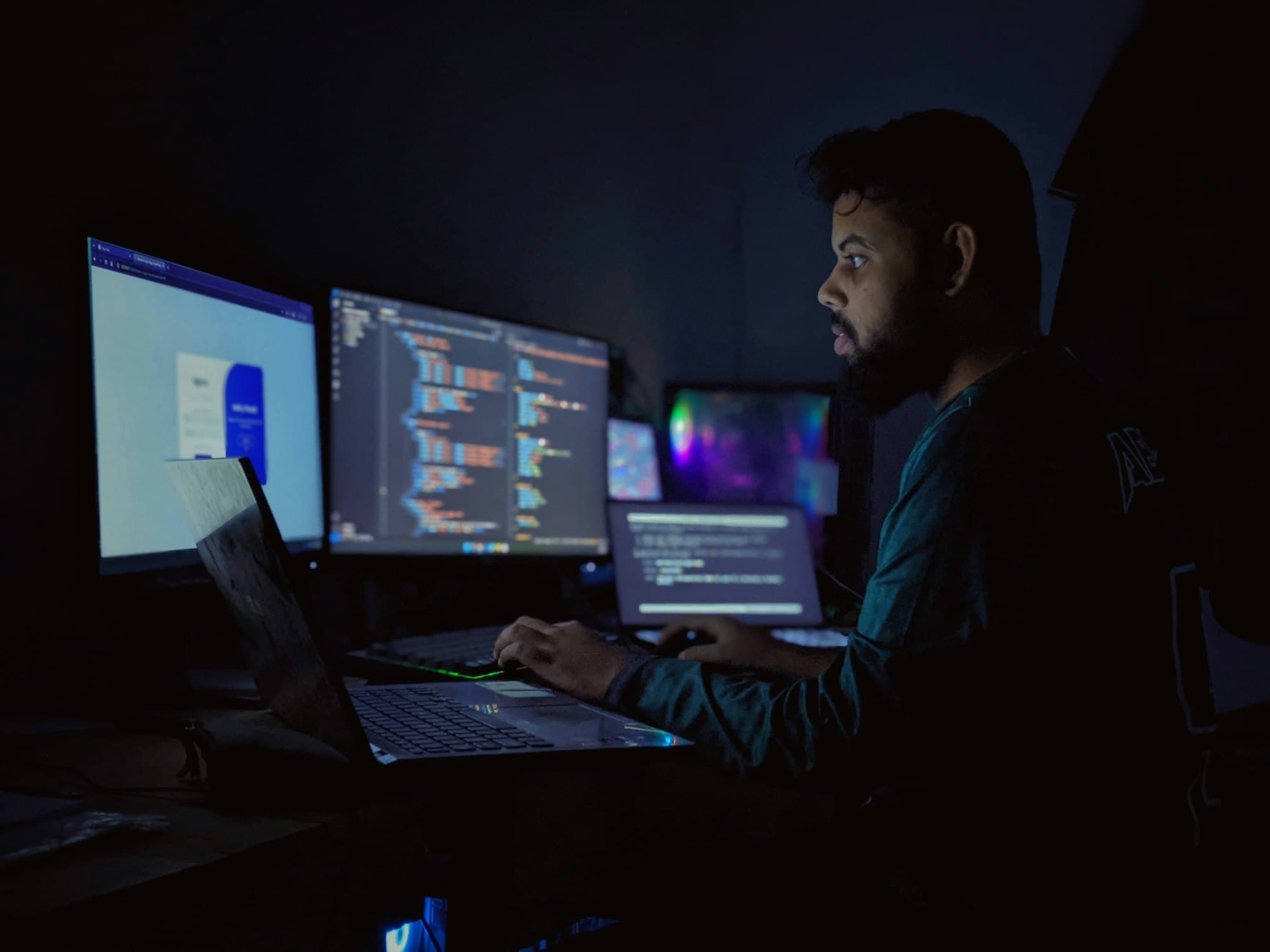 A developer types at a laptop in a dark room lit by monitors full of out-of-focus code